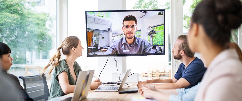 Vier Kollegen an einem Tisch in einer Videokonferenz mit einem zugeschalteten Kollegen
